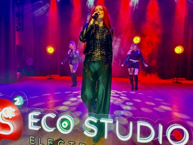 Trzy kobiety występują na scenie. W centrum stoi śpiewająca kobieta z długimi czerwonymi włosami trzymająca mikrofon i ubrana w czarny skórzany strój. Za nią dwie tancerki w ciemnych strojach. Scena oświetlona jest czerwonym i fioletowym światłem z dymem w tle, a na pierwszym planie widoczny jest neonowy napis przy krawędzi sceny.