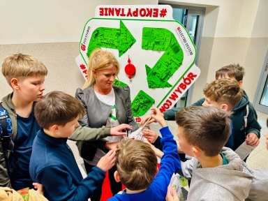 Kobieta stoi pośrodku grupy siedmiu chłopców w jasnym korytarzu. Chłopcy sięgają po ulotki trzymane przez kobietę przed dużym białym stojakiem z zielonym symbolem recyklingu i czerwonymi akcentami.