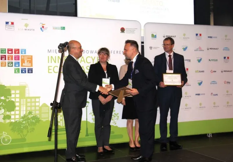 Cztery osoby na scenie podczas wręczenia nagród na konferencji, dwie osoby witają się i wymieniają dokumentami.