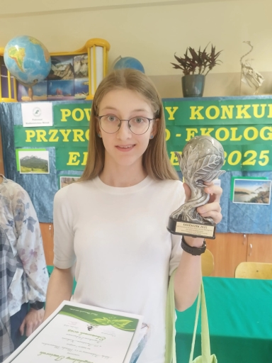 Jasna sala lekcyjna. Dziewczyna w białej bluzce i okularach stoi w sali. Trzyma srebrny puchar i dyplom. Za nią zielono-żółty baner z napisem dotyczącym konkursu przyrodniczo-ekologicznego. W tle globus i roślina doniczkowa.