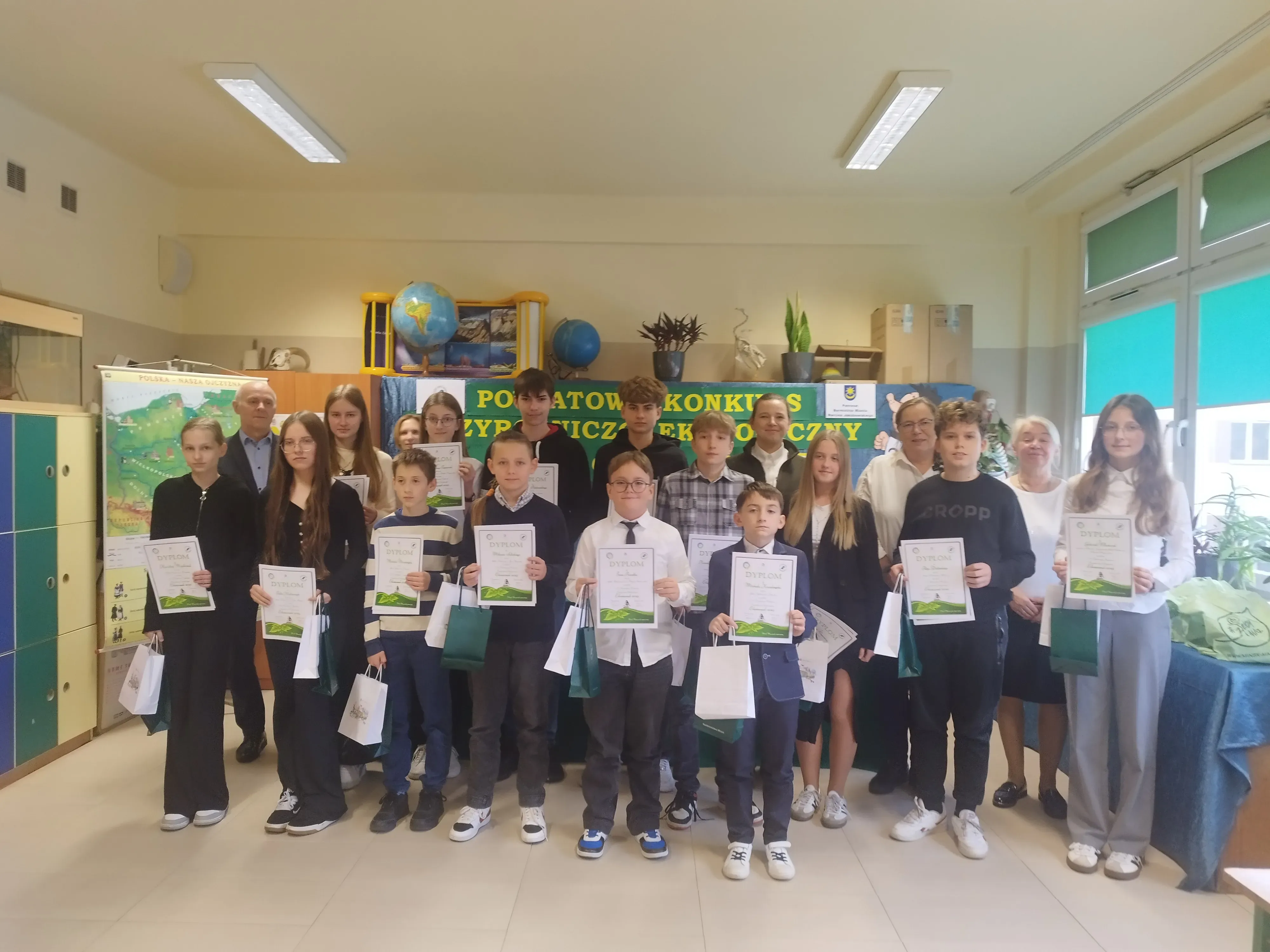 Grupa głównie dzieci i nastolatków, stoi w klasie szkolnej. Każda osoba trzyma dyplom i torbę upominkową. W tle zielony plakat z napisem konkursu oraz mapa i globus na szafkach. Pomieszczenie jest jasno oświetlone, widoczne okna z zielonymi roletami po prawej stronie.