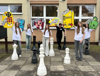 Siedem osób stoi na zewnątrz przed beżowym budynkiem z oknami. Większość nosi białe koszulki, jedna osoba ma czarną. Każda trzyma dużą, kolorową wyciętkę z rysunkową postacią przypominającą urządzenia elektroniczne z oczami. Przed nimi na bruku ustawione są duże czarne i białe figury szachowe.