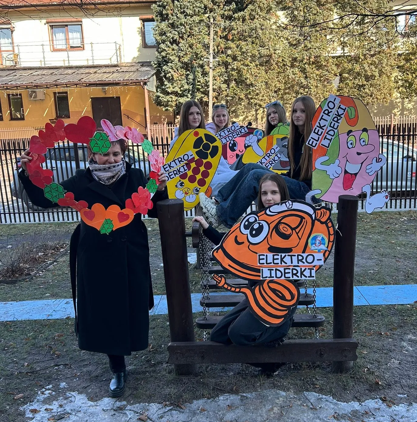 Sześć osób stoi i siedzi na drewnianej huśtawce na zewnątrz przed metalowym płotem. Osoby trzymają kolorowe kartonowe postacie i papierowy wieniec w kształcie serca. W tle widać budynek i drzewa, ziemia z resztkami śniegu.