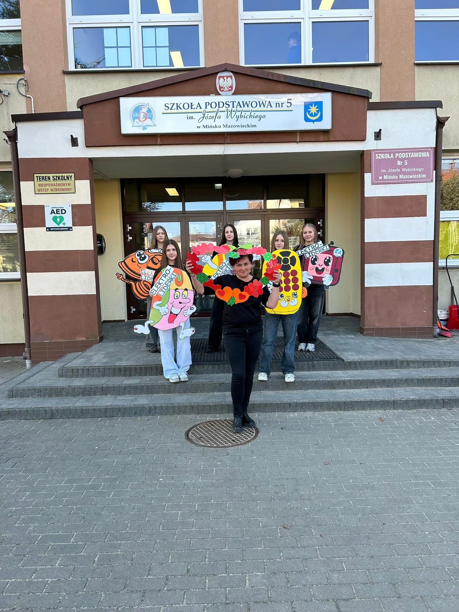 Sześć osób stoi na schodach przed wejściem do szkoły. Jedna kobieta na pierwszym planie trzyma wieniec z czerwonych papierowych serc, pozostałe osoby trzymają kolorowe kartonowe postacie i znaki. Budynek ma jasnobrązową elewację i duże okna.