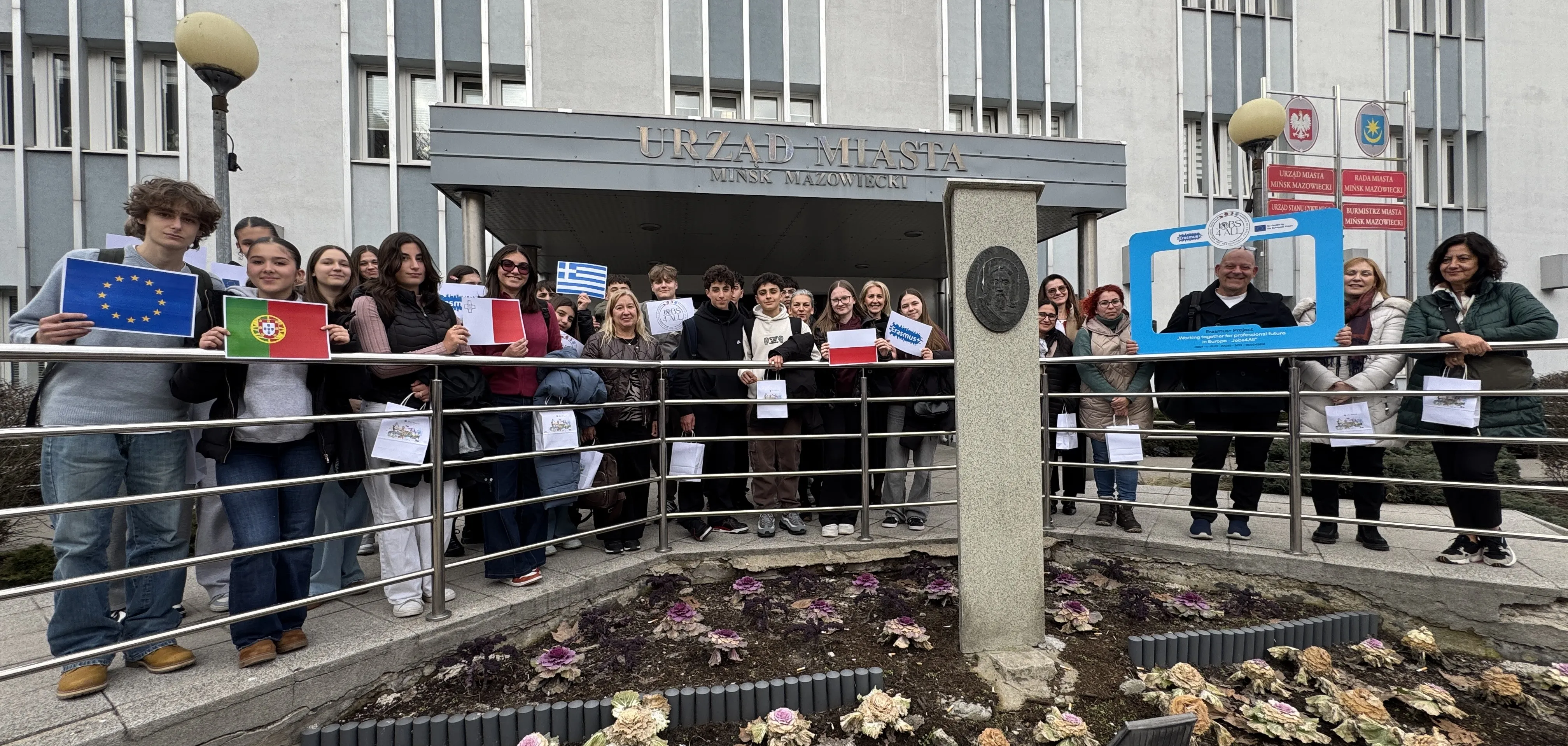 Około siedemnastu nastolatków i trzy dorosłe osoby siedzą i rozmawiają wokół dużego drewnianego stołu w jasnej sali konferencyjnej. Po prawej stoi kobieta przy ściennym monitorze z kolorowym slajdem, za grupą stoi flipchart i okno z pionowymi żaluzjami.