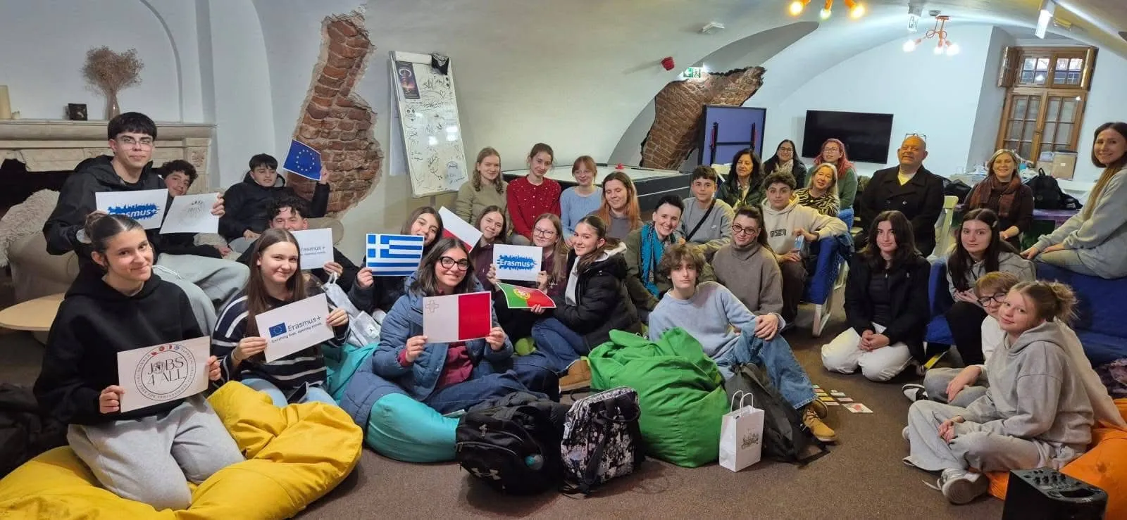 Około trzydziestu osób siedzi w sali z masywnym, białym, sklepionym sufitem i odsłoniętymi fragmentami ceglanej ściany. Młodzi ludzie siedzą na kolorowych workach sako i krzesłach, trzymają małe flagi i plakaty z logo Erasmus i flagami Grecji, Malty, Portugalii oraz Unii Europejskiej. W tle znajduje się kominek, tablica suchościeralna, telewizor i okno. Sala jest oświetlona sztucznym światłem.