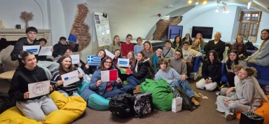 Około trzydziestu osób siedzi w sali z masywnym, białym, sklepionym sufitem i odsłoniętymi fragmentami ceglanej ściany. Młodzi ludzie siedzą na kolorowych workach sako i krzesłach, trzymają małe flagi i plakaty z logo Erasmus i flagami Grecji, Malty, Portugalii oraz Unii Europejskiej. W tle znajduje się kominek, tablica suchościeralna, telewizor i okno. Sala jest oświetlona sztucznym światłem.