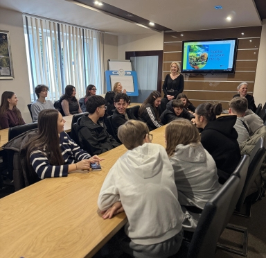 Około piętnastu młodych osób siedzi przy długim drewnianym stole w sali konferencyjnej. Dwie dorosłe kobiety stoją i siedzą przy ścianie z ekranem pokazującym kolorowy slajd. W pomieszczeniu są duże pionowe rolety, jasne oświetlenie sufitowe i czarne krzesła.