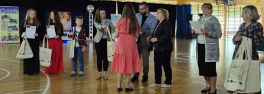 Grupa ośmiu osób w hali sportowej przyjmuje i wręcza torby płócienne oraz dyplomy, dwie młode kobiety po lewej trzymają dyplomy i torby, chłopiec i kobieta obok z torbami, kobieta w różowej sukience stoi tyłem, trzy osoby po prawej przygotowane do wręczenia dokumentów i toreb.