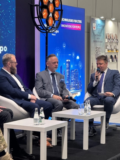 Trzech mężczyzn w garniturach siedzi na białych fotelach w pomieszczeniu konferencyjnym. Dwóch mężczyzn trzyma mikrofony, a jeden z nich mówi. Przed nimi stoją dwie białe stoliki z butelkami wody i szklankami. W tle niebieski ekran z grafiką przedstawiającą wieżowce i napisem dotyczącym technologii dla miast oraz baner LiftExpo Poland.
