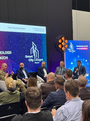 Pięciu mężczyzn siedzi na scenie podczas panelu dyskusyjnego na targach technologii miejskich. Czterech ma garnitury, jeden mundur wojskowy. Przed nimi siedzi publiczność. W tle duże ekrany z grafiką i nazwą wydarzenia Smart City Expo Poland, datą i miejscem oraz napisem Technologies for Cities. Scena oświetlona, ciemne tło.