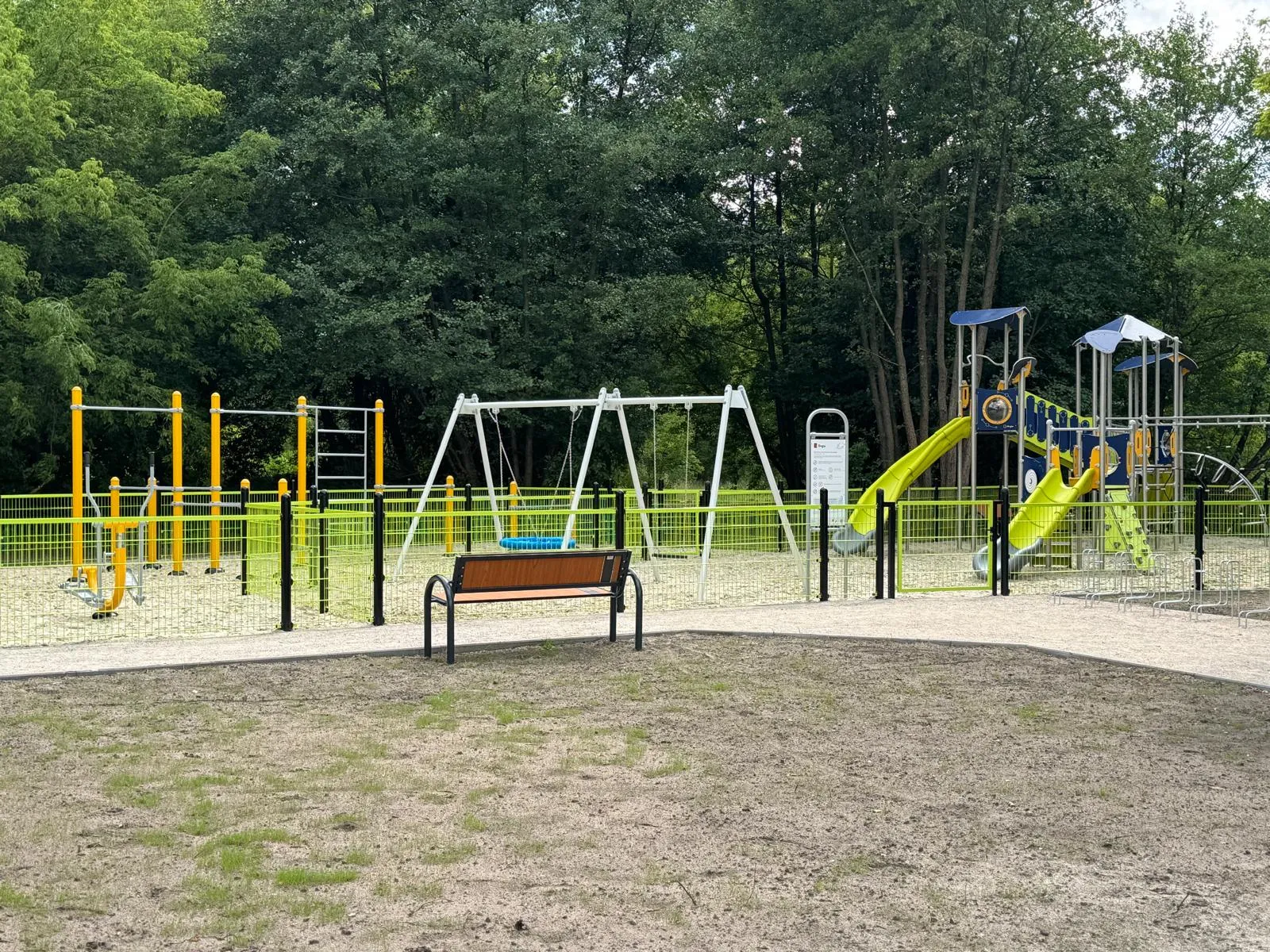 Ogrodzony plac zabaw z huśtawkami, zjeżdżalniami i sprzętem do wspinaczki na tle drzew.