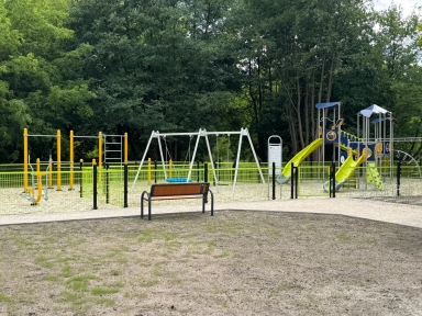 Ogrodzony plac zabaw z huśtawkami, zjeżdżalniami i sprzętem do wspinaczki na tle drzew.