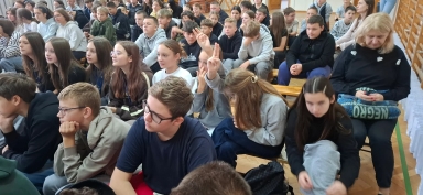 Duża grupa młodych osób siedzi na drewnianych ławkach w sali z drewnianymi panelami na ścianie, większość patrzy przed siebie, dwie dziewczyny wykonują znak zwycięstwa rękami, kobieta siedząca po prawej trzyma torbę na kolanach i patrzy w dół.