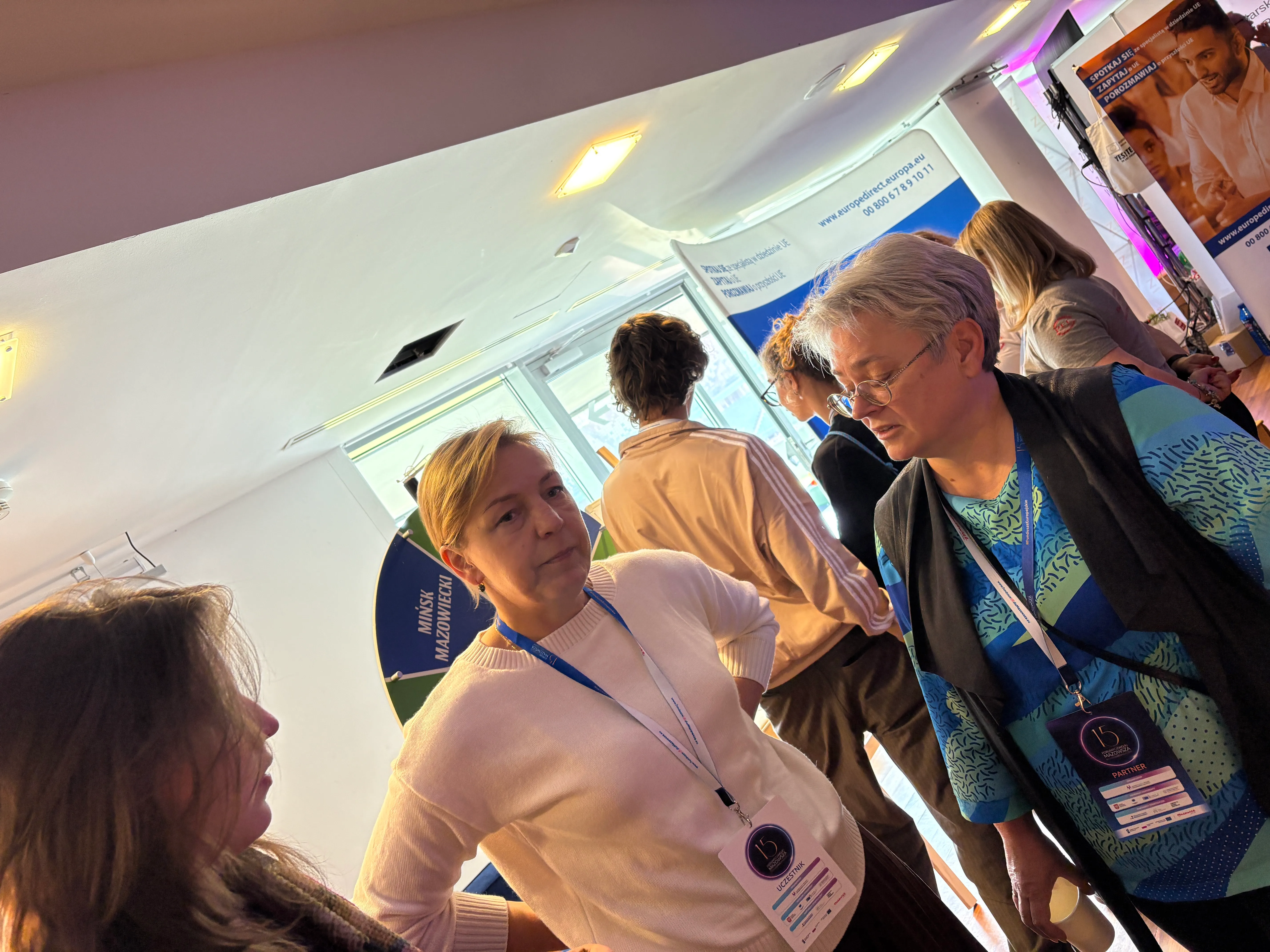 Trzy kobiety rozmawiają w jasno oświetlonym, zamkniętym pomieszczeniu podczas wydarzenia. Dwie z nich stoją przodem do siebie, trzecia jest częściowo zwrócona bokiem. Wszystkie mają przypięte identyfikatory na niebieskich smyczach. W tle stoi kilka osób i widoczne są plakaty z tekstem oraz koło z napisem „MIŃSK MAZOWIECKI”.