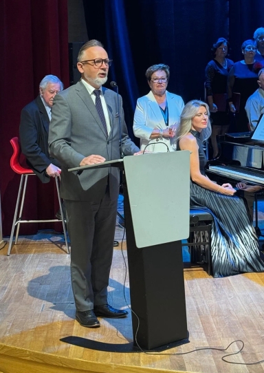 Mężczyzna w garniturze przemawia przy mównicy na scenie. Za nim stoi kobieta w białej marynarce i kilka innych osób. Kobieta w długiej sukni siedzi przy fortepianie. W tle ciemna kurtyna i piętnastolitrowa podłoga.