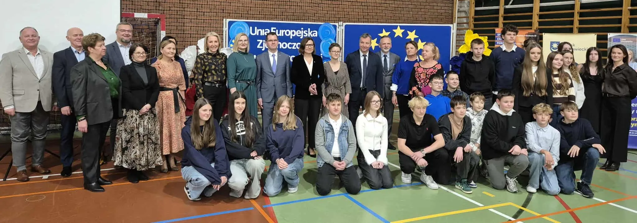 Grupa około 35 osób, w tym dorośli i młodzież, stoi i klęczy na hali sportowej przed planszami z symbolami Unii Europejskiej.