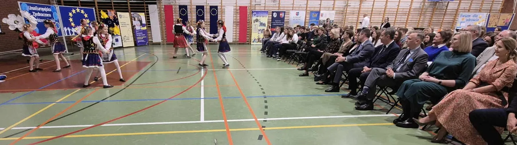 Grupa dzieci w strojach ludowych tańczy na hali sportowej, po prawej stronie siedzi publiczność oglądająca występ, w tle wiszą flagi Polski i Unii Europejskiej oraz stoją banery informacyjne.