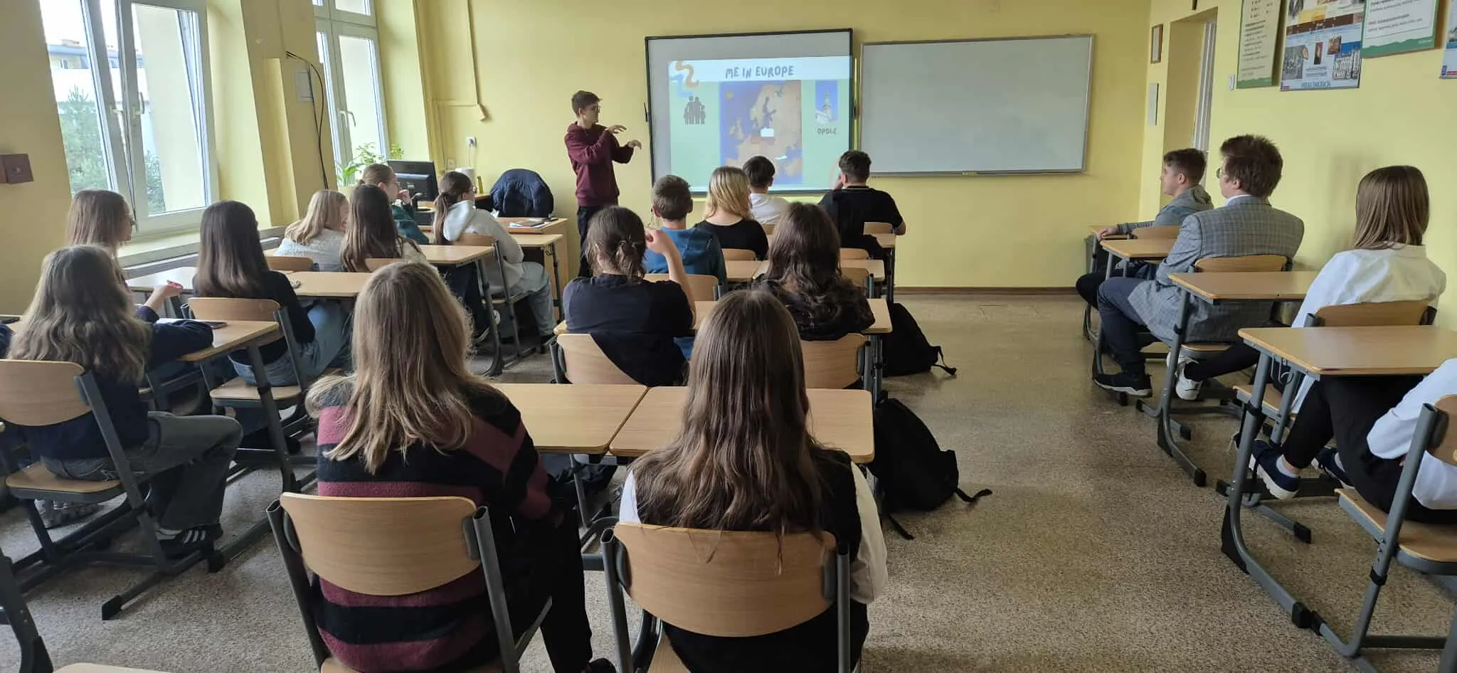 Uczniowie siedzą w klasie na krzesłach przy biurkach zwróceni w stronę tablicy interaktywnej, na której chłopiec prezentuje mapę Europy. Klasa ma żółte ściany i duże okna po lewej stronie.