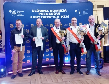 Pięciu mężczyzn stoi na tle niebieskiego baneru z napisem „Główna Komisja Pojazdów Zabytkowych PZM”. Trzech z nich trzyma puchary i ma na sobie czerwone szarfy, dwaj pozostali trzymają dyplomy. Wszyscy ubrani są w półformalne stroje. W tle znajduje się wnętrze budynku z oświetleniem sceny.