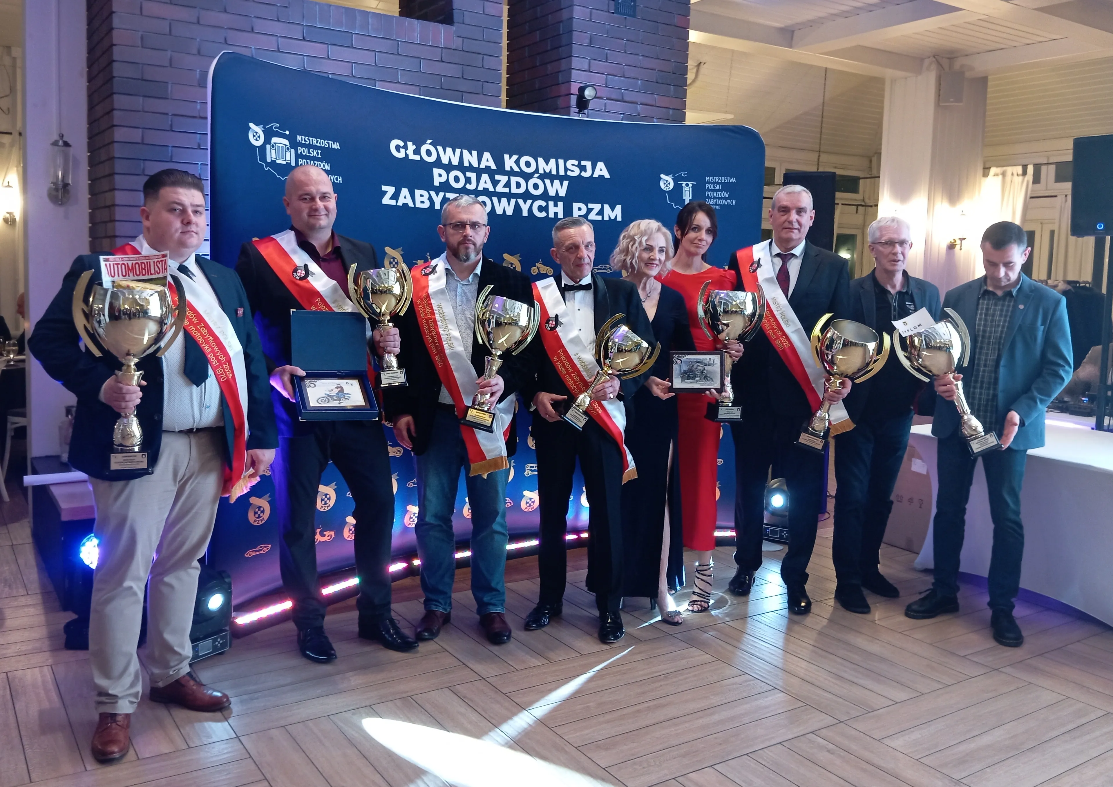 Dziewięć osób stoi przed niebieskim banerem z białym napisem, każda trzyma puchar lub nagrodę, siedem osób ma na sobie czerwono-białe szarfy, wnętrze dobrze oświetlone, tło z ceglaną ścianą i drewnianą podłogą.