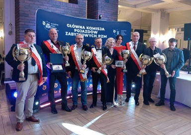 Dziewięć osób stoi przed niebieskim banerem z białym napisem, każda trzyma puchar lub nagrodę, siedem osób ma na sobie czerwono-białe szarfy, wnętrze dobrze oświetlone, tło z ceglaną ścianą i drewnianą podłogą.