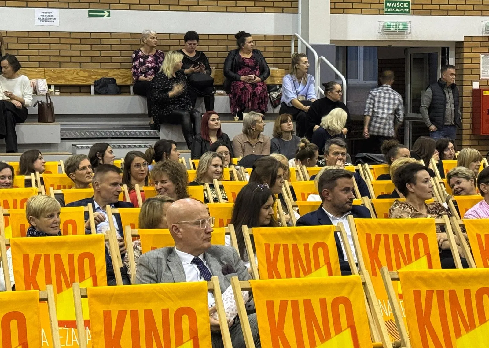 Wnętrze hali z dużą grupą dorosłych osób siedzących na żółtych, składanych krzesłach oznaczonych napisem „KINO”. W tle kilka osób siedzi na drewnianych ławkach przy ścianie z cegieł. Na ścianie widoczne zielone oznaczenie wyjścia ewakuacyjnego.