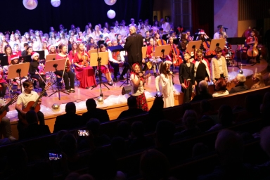 Grupa dzieci i młodzieży na scenie podczas koncertu, część z nich gra na instrumentach smyczkowych i gitarach, inne śpiewają lub recytują, widzowie siedzą na ciemnej widowni, scenę dekorują białe ozdoby i wiszące kule, atmosfera świąteczna.