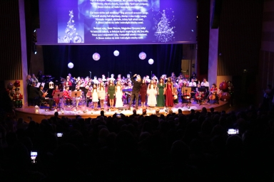 Grupa młodych kobiet śpiewa na scenie teatru z towarzyszeniem orkiestry. Na tle czarnej kurtyny wiszą białe dekoracje w kształcie bombek. Scena jest oświetlona, publiczność siedzi w ciemności. Nad sceną wyświetlany jest tekst z motywem świątecznym.