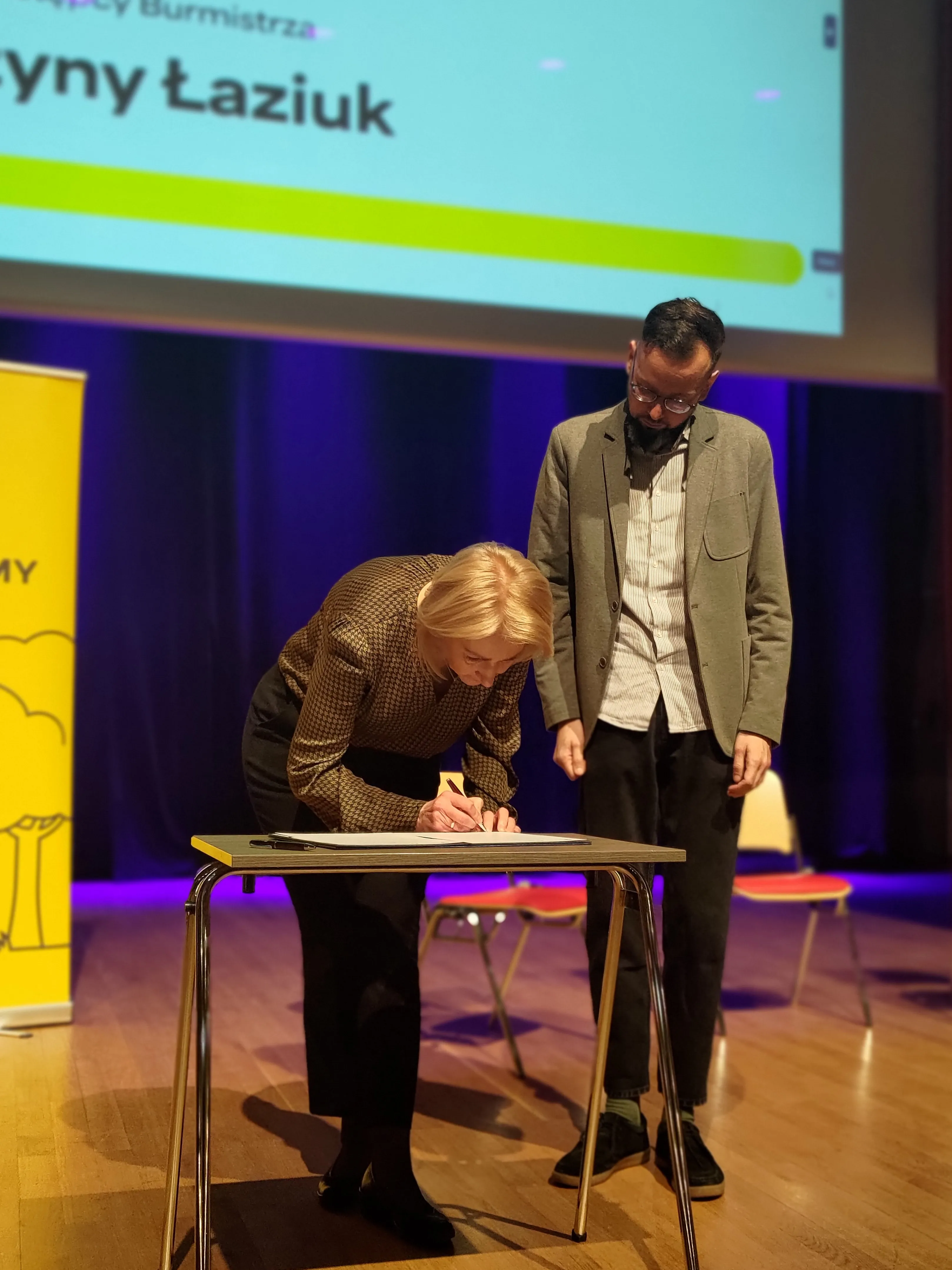 Kobieta pochyla się nad stolikiem i podpisuje dokument. Mężczyzna stoi obok niej i patrzy w dół. Scena jest na scenie z fioletowym tłem, drewnianą podłogą i krzesłami w tle. Po lewej stoi żółty pionowy baner, u góry widoczny fragment projekcji.