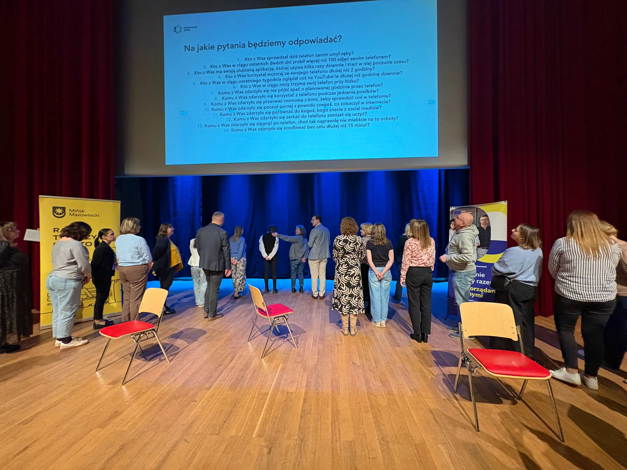 Około osiemnastu osób stoi na drewnianej scenie w sali konferencyjnej zwróconych twarzami do dużego, jasnego ekranu projekcyjnego. Scena ma niebieskie zasłony w tle i czerwone teatralne kotary po bokach. Po lewej i prawej stoją żółte słupki reklamowe. Na scenie stoją rozrzucone krzesła z czerwonymi siedziskami.