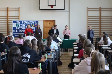 Sala gimnastyczna z ustawionymi rzędami biurek, przy których siedzi grupa uczniów. Przód sali zajmuje pięć kobiet, jedna z nich mówi do mikrofonu. Na ścianie za nimi wisi tablica z napisem dotyczącym konkursu ortograficznego. Na ścianach drewniane drabinki gimnastyczne, pod sufitem kosz do koszykówki.