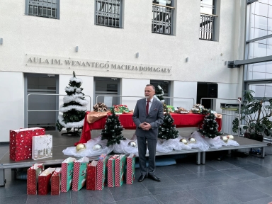Mężczyzna w garniturze stoi obok sceny udekorowanej sztucznymi choinkami, białą tkaniną i dużymi złotymi oraz srebrnymi bombkami. Na scenie znajduje się stół z czerwonym obrusem i dekoracjami. Przed sceną stoi kilka kolorowych toreb prezentowych. W tle na ścianie widoczny jest napis "AULA IM. WENANTEGO MACIEJA DOMAGAŁY". Wnętrze jest jasne, z dużymi oknami po prawej stronie.