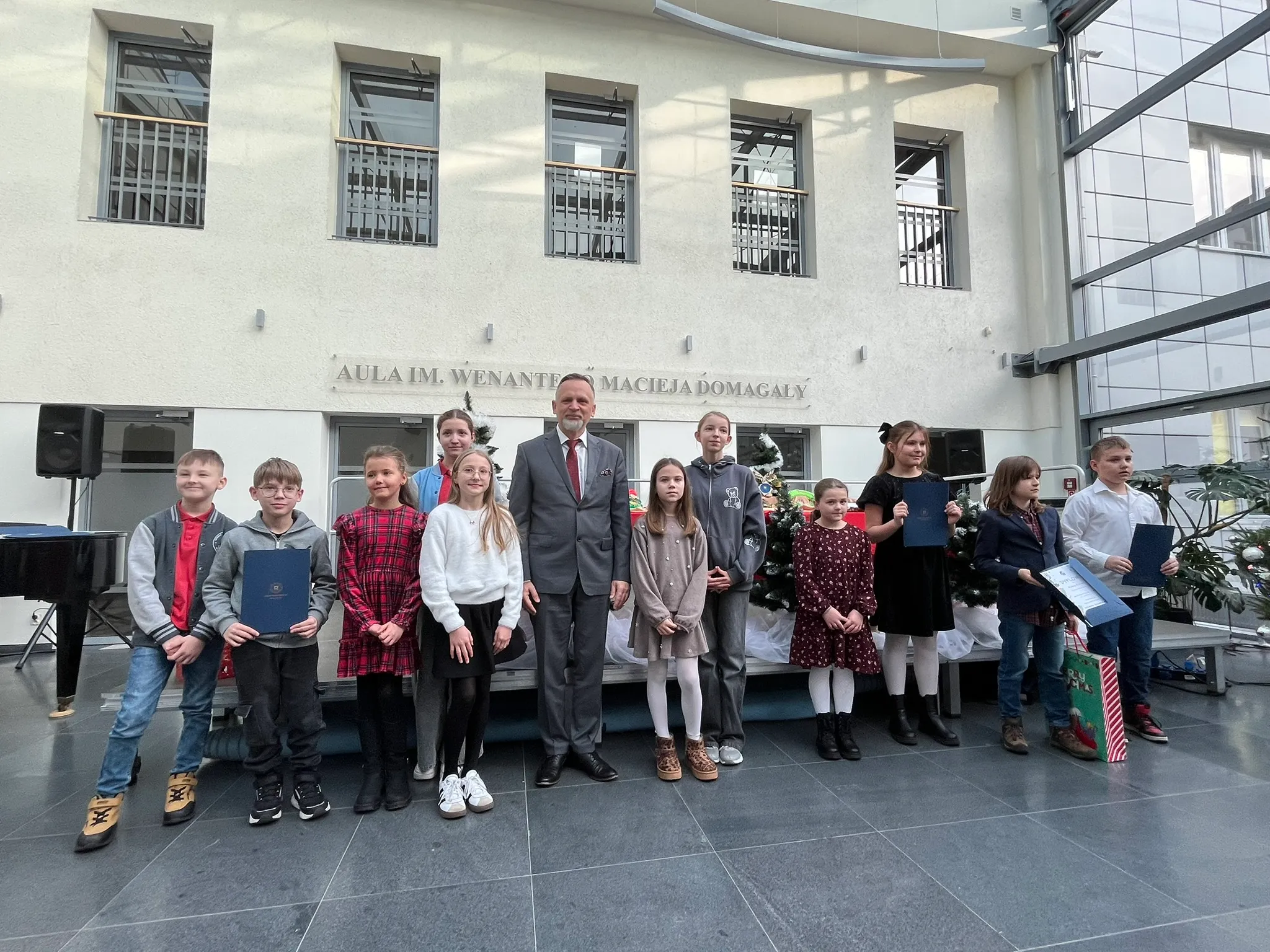 Grupa jedenastu dzieci i dorosły mężczyzna w garniturze stoi w jasnym holu przed ścianą z napisem „AULA IM. WENANTEGO MACIEJA DOMAGAŁY”. Dzieci trzymają teczki i prezenty. Za nimi stoją udekorowane choinki. Pomieszczenie ma duże okna i szare podłogi.