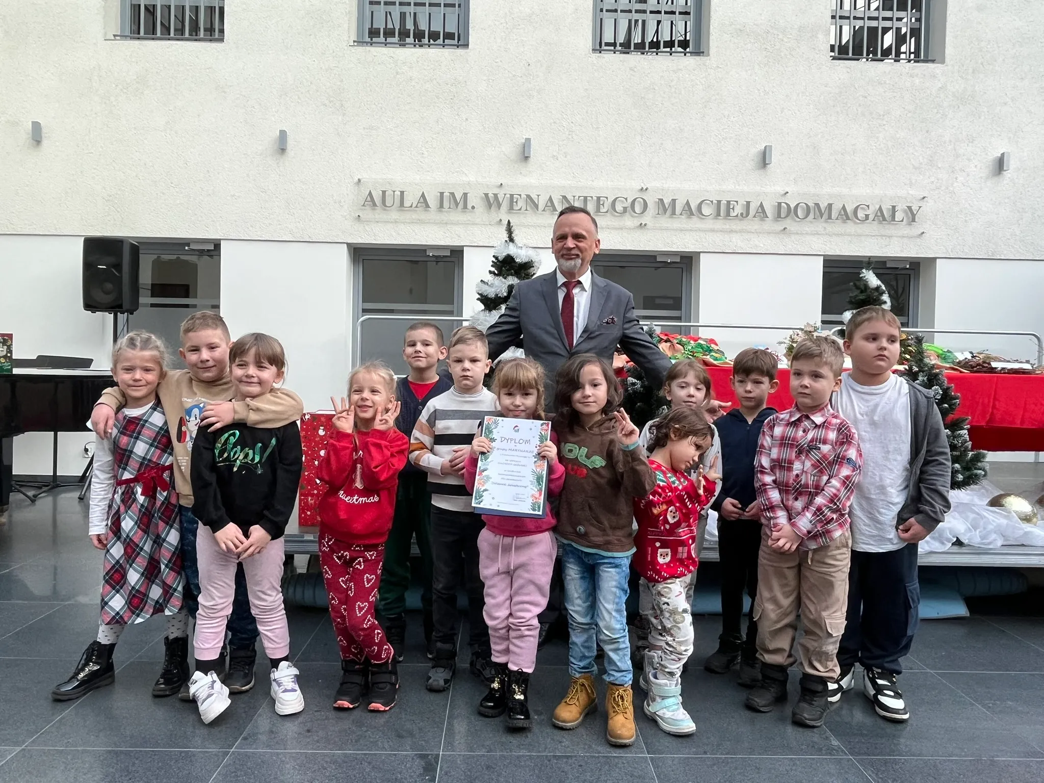 Mężczyzna w garniturze stoi za grupą dwunastu dzieci na tle ściany z napisem, w pomieszczeniu o jasnych ścianach. Dzieci ubrane są w kolorowe ubrania, jedna trzyma dyplom. W tle stoją udekorowane choinki i stół z czerwonym obrusem.