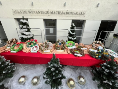 Stoisko z ozdobnymi wiankami na tle ściany z napisem „AULA IM. WENANTEGO MACIEJA DOMAGAŁY”, ozdobione dwoma choinkami z białą lametą, na stole czerwony i złoty materiał, pod stołem sztuczny śnieg i cztery srebrne jajka, w tle drzwi i metalowe barierki.
