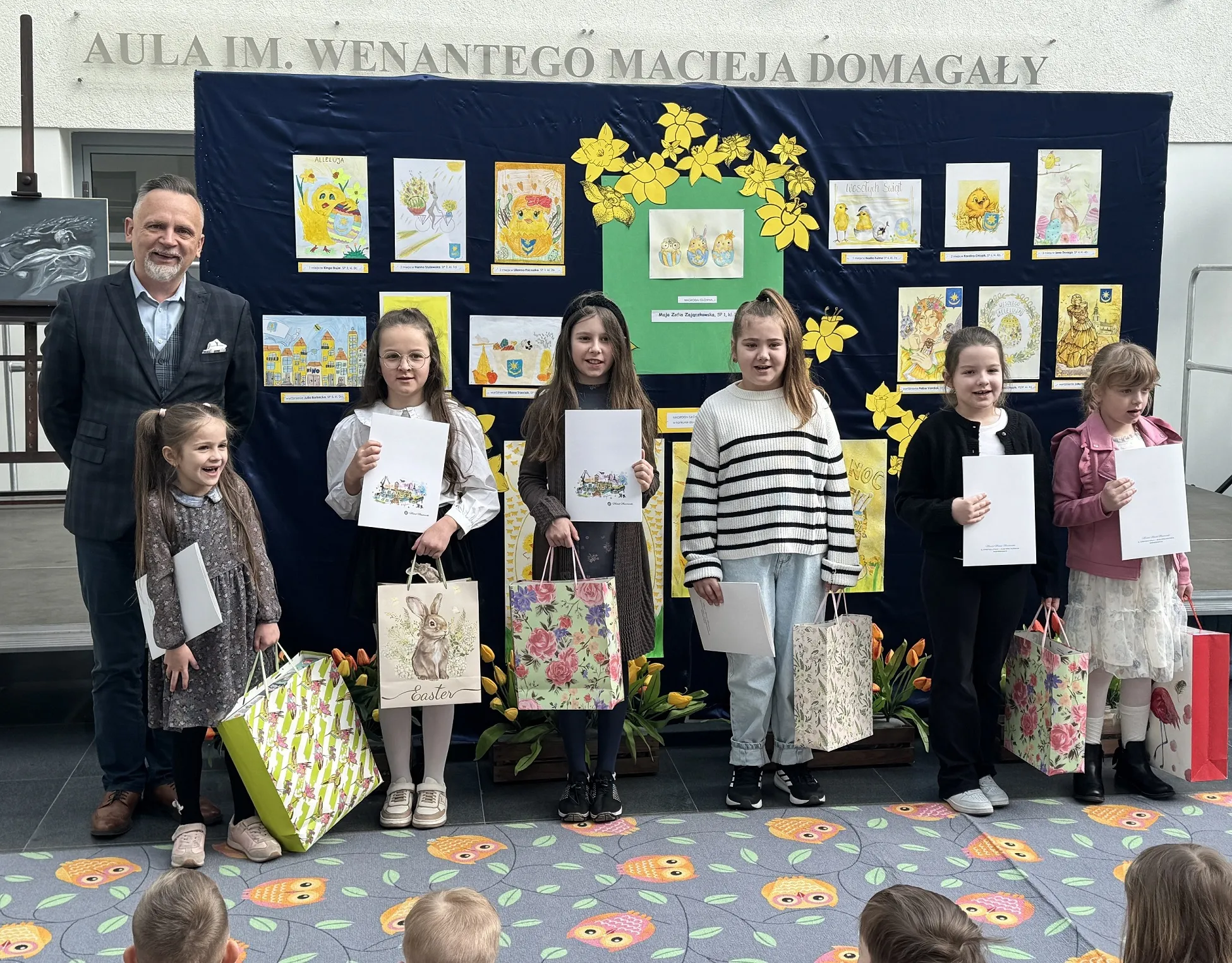 Mężczyzna i sześć dziewczynek stoją w rzędzie przed granatową tablicą z kolorowymi rysunkami. Dzieci trzymają białe dyplomy i ozdobne torebki prezentowe. Scena odbywa się wewnątrz, na podłodze widoczny jest dywan z wzorem sów. Za tablicą widoczne żółte dekoracje kwiatowe.