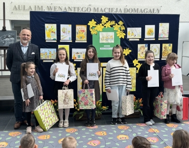 Mężczyzna i sześć dziewczynek stoją w rzędzie przed granatową tablicą z kolorowymi rysunkami. Dzieci trzymają białe dyplomy i ozdobne torebki prezentowe. Scena odbywa się wewnątrz, na podłodze widoczny jest dywan z wzorem sów. Za tablicą widoczne żółte dekoracje kwiatowe.