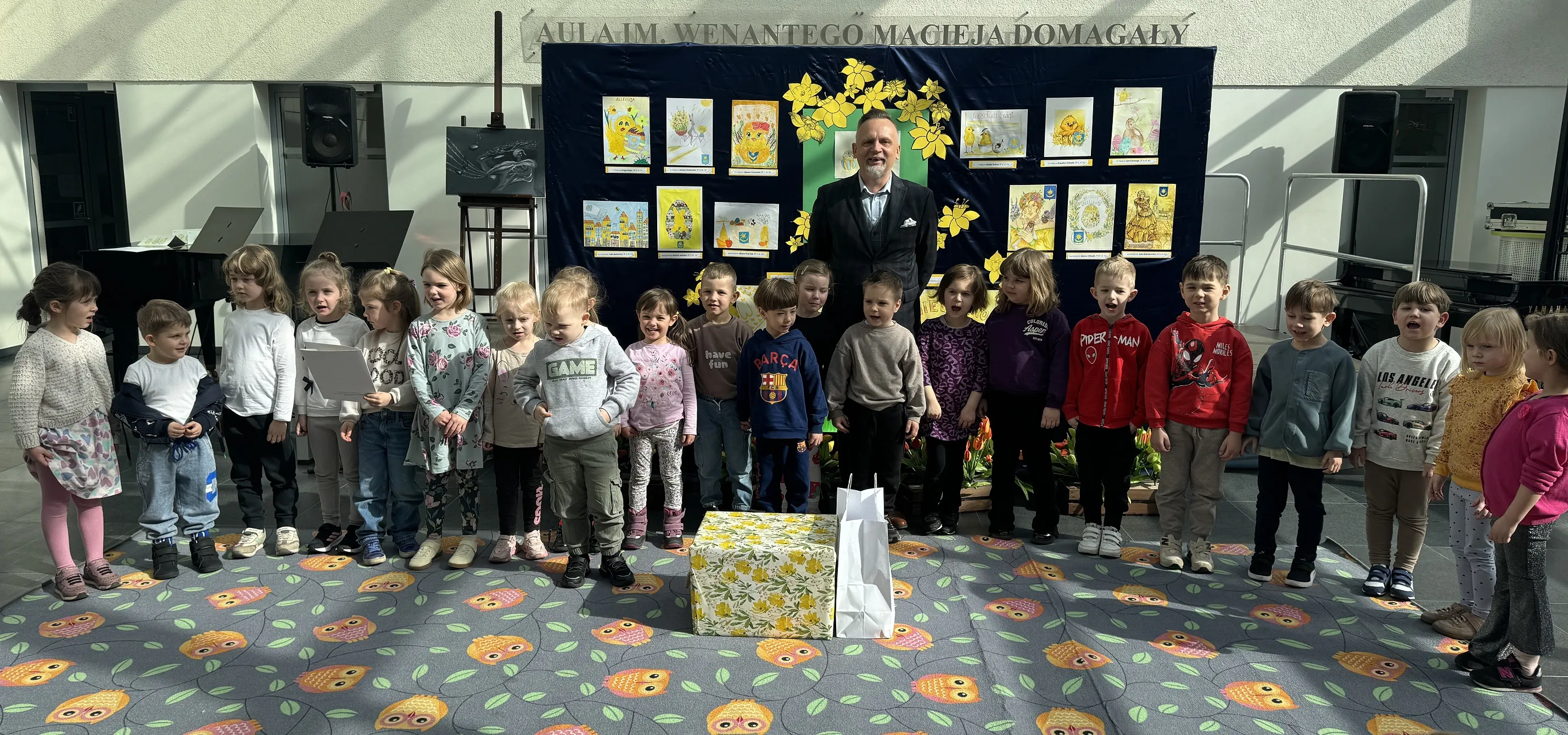 Grupa około 22 dzieci stoi w jednym rzędzie na wewnętrznym, dobrze oświetlonym holu. Jeden dorosły mężczyzna stoi za dziećmi przy ciemnej tablicy z rysunkami i żółtymi dekoracjami. Na podłodze kolorowy dywan z wzorem sów. Przed grupą leży duże pudełko w kwiatowy wzór i biała torba.