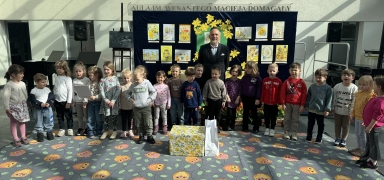 Grupa około 22 dzieci stoi w jednym rzędzie na wewnętrznym, dobrze oświetlonym holu. Jeden dorosły mężczyzna stoi za dziećmi przy ciemnej tablicy z rysunkami i żółtymi dekoracjami. Na podłodze kolorowy dywan z wzorem sów. Przed grupą leży duże pudełko w kwiatowy wzór i biała torba.