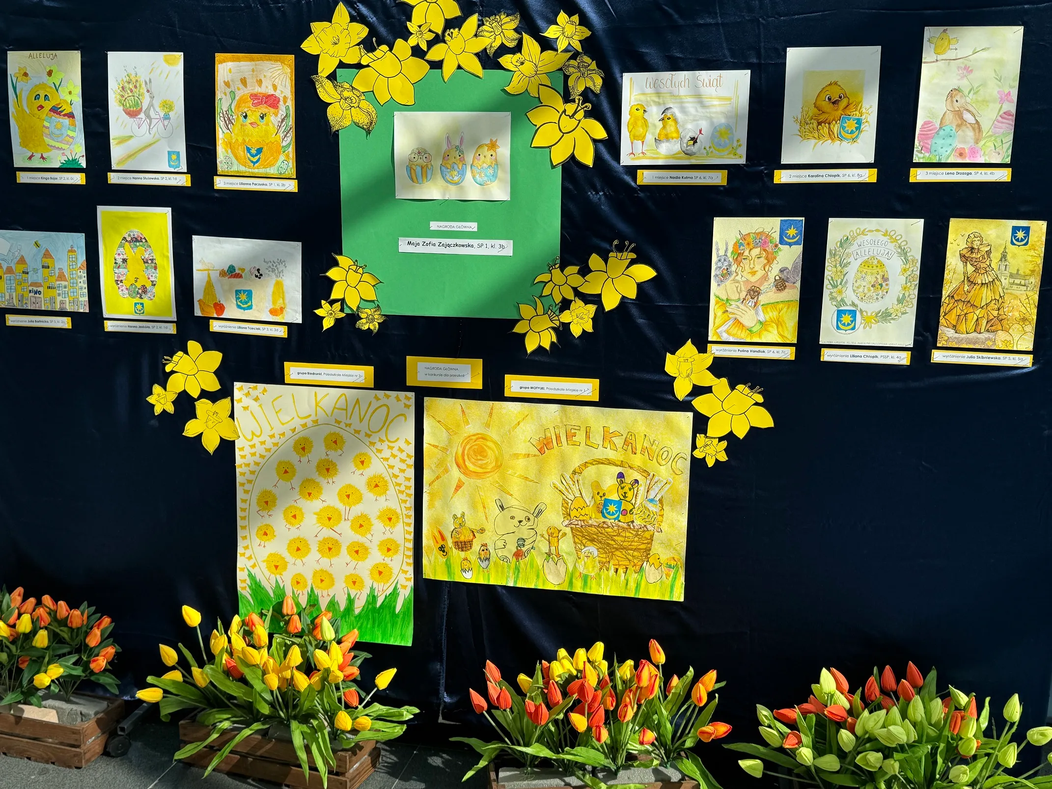 Kilkanaście wielkanocnych rysunków i pocztówek ułożonych na ciemnym tle.  
Dwa duże centralne plakaty w żółto-zielonej kolorystyce z napisem WIELKANOC.  
Żółte wycinanki kwiatów (żonkile) rozmieszczone wokół centralnej części.  
Kilka drewnianych skrzynek z żółto-pomarańczowo-czerwonymi tulipanami u dołu.