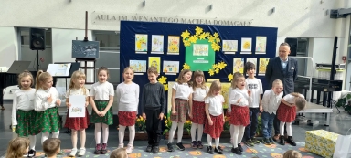 Grupa dzieci stoi w rzędzie na wewnętrznej scenie przed granatową tablicą z kolorowymi rysunkami i żółtymi kwiatami. Dzieci noszą białe bluzki i czerwono-zielone spódniczki lub ciemne spodnie. Po prawej stoi mężczyzna w marynarce. Za nimi widać jasną ścianę z napisem i instrumenty muzyczne. Tulipany i dekoracyjne pudełka stoją na podłodze przed sceną.