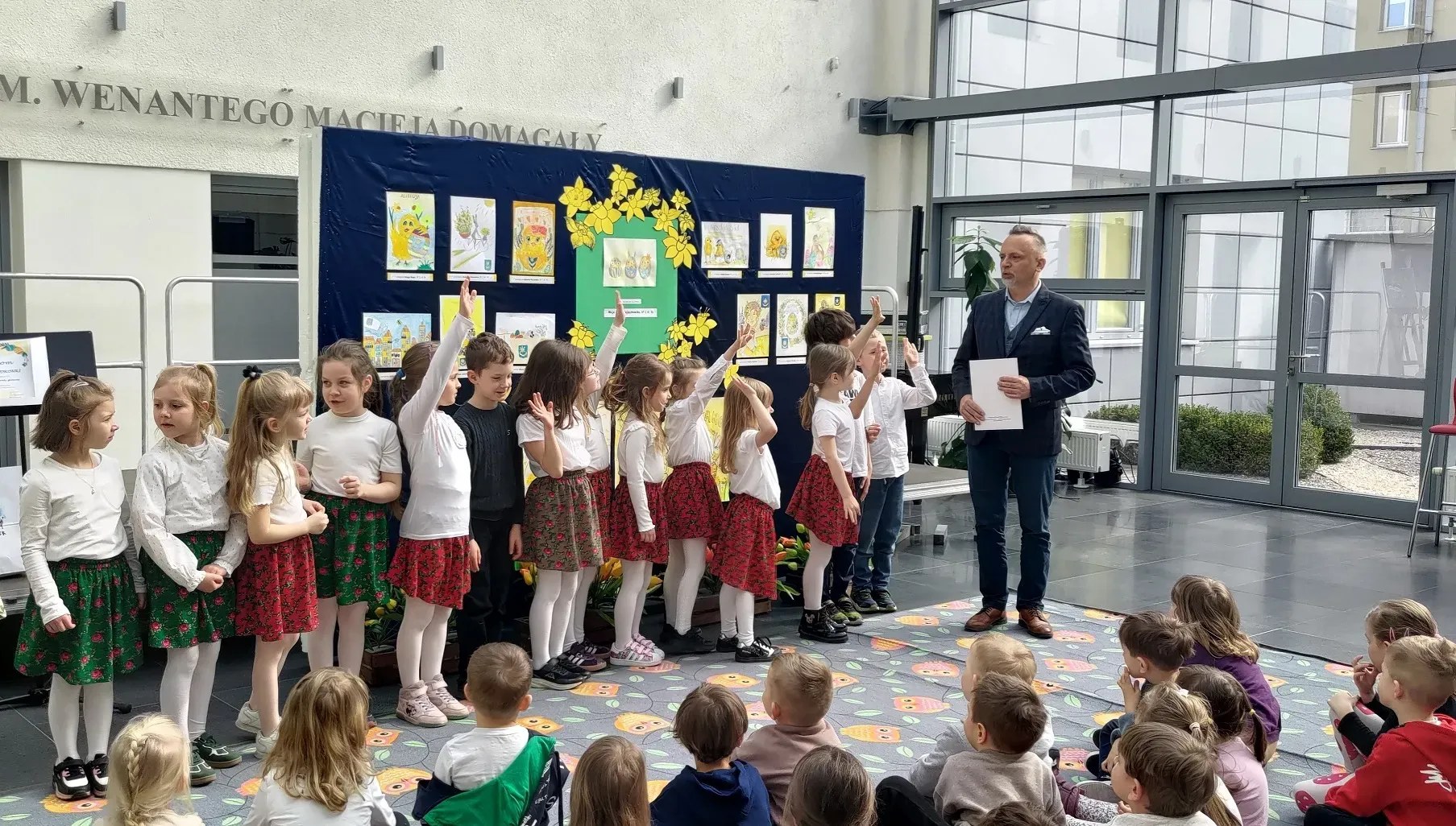 Grupa dzieci stoi w rzędzie przed granatową tablicą z rysunkami i dekoracją z żółtych kwiatów. Dzieci noszą białe bluzki i czerwono-zielone spódniczki lub białe koszule. Mężczyzna w marynarce trzyma kartki po prawej stronie. Inne dzieci siedzą na kolorowym dywanie.