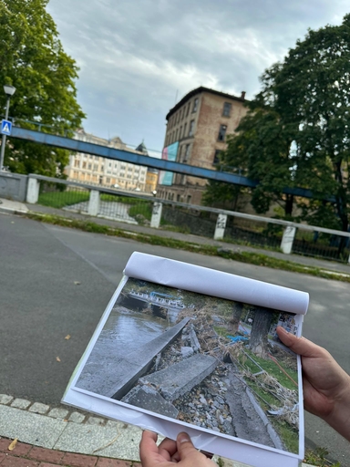 Dwie dłonie trzymają zrolowane i rozłożone zdjęcie z widokiem na brzeg rzeki. W tle na zewnątrz widoczny niebieski most nad kanałem, stary ceglany budynek i duże drzewa. Niebo jest zachmurzone, jest dzień.