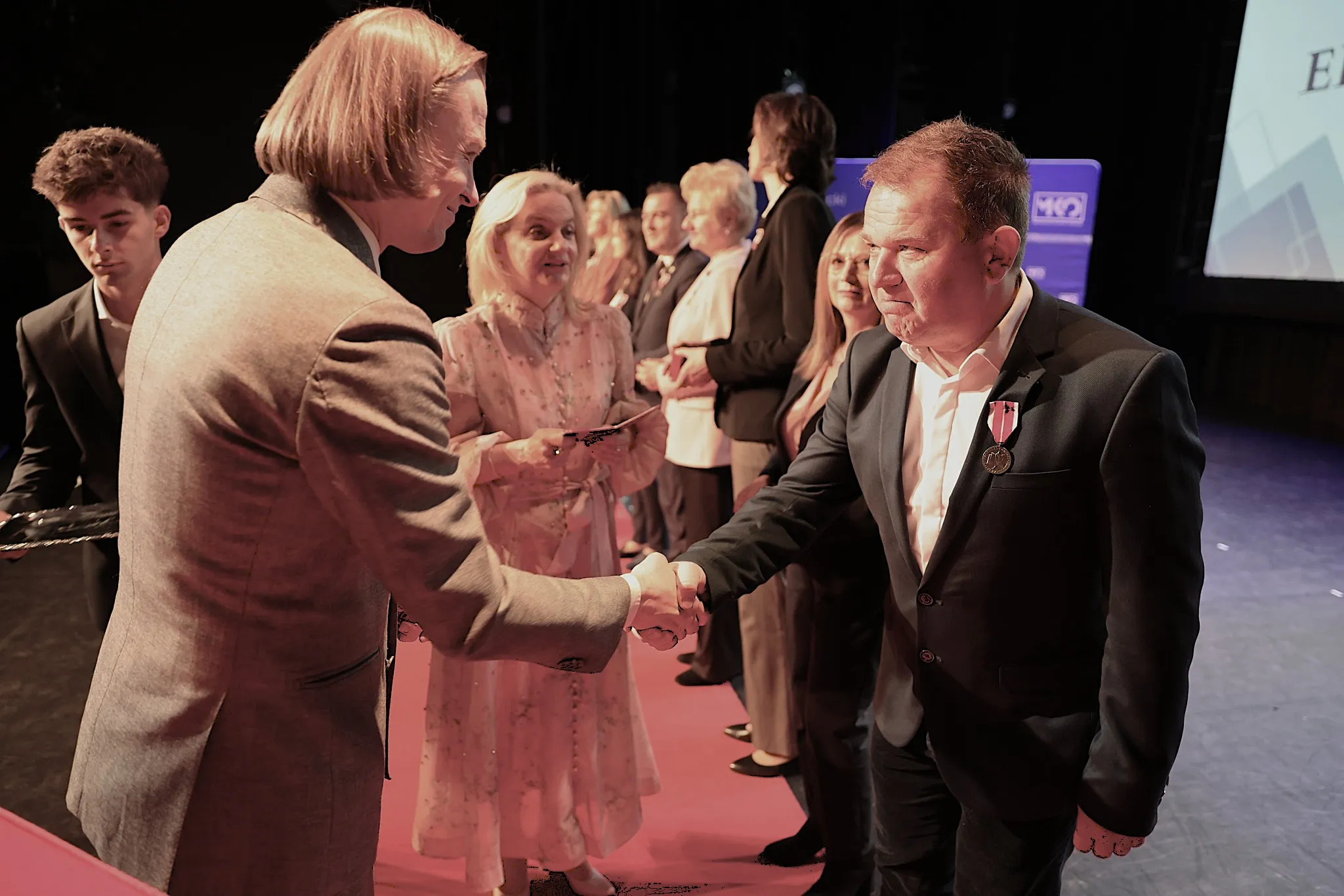 Dwaj mężczyźni ściskają sobie ręce na scenie podczas uroczystości. Mężczyzna po prawej ma medal na marynarce. W tle stoi kilku uśmiechniętych ludzi ustawionych w rzędzie. Otoczenie jest ciemne, scena oświetlona.