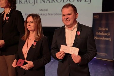 Usmiechnięty mężćzyzna i kobieta stoją na scenie podczas wręczenia medali. Oboje mają przypięte medale do ciemnych marynarek. Mężczyzna po prawej trzyma otwarty dyplom. W tle widoczny jest baner z napisem „Dzień Edukacji Narodowej”. Scena jest ciemna, a oświetlenie skupia się na osobach.