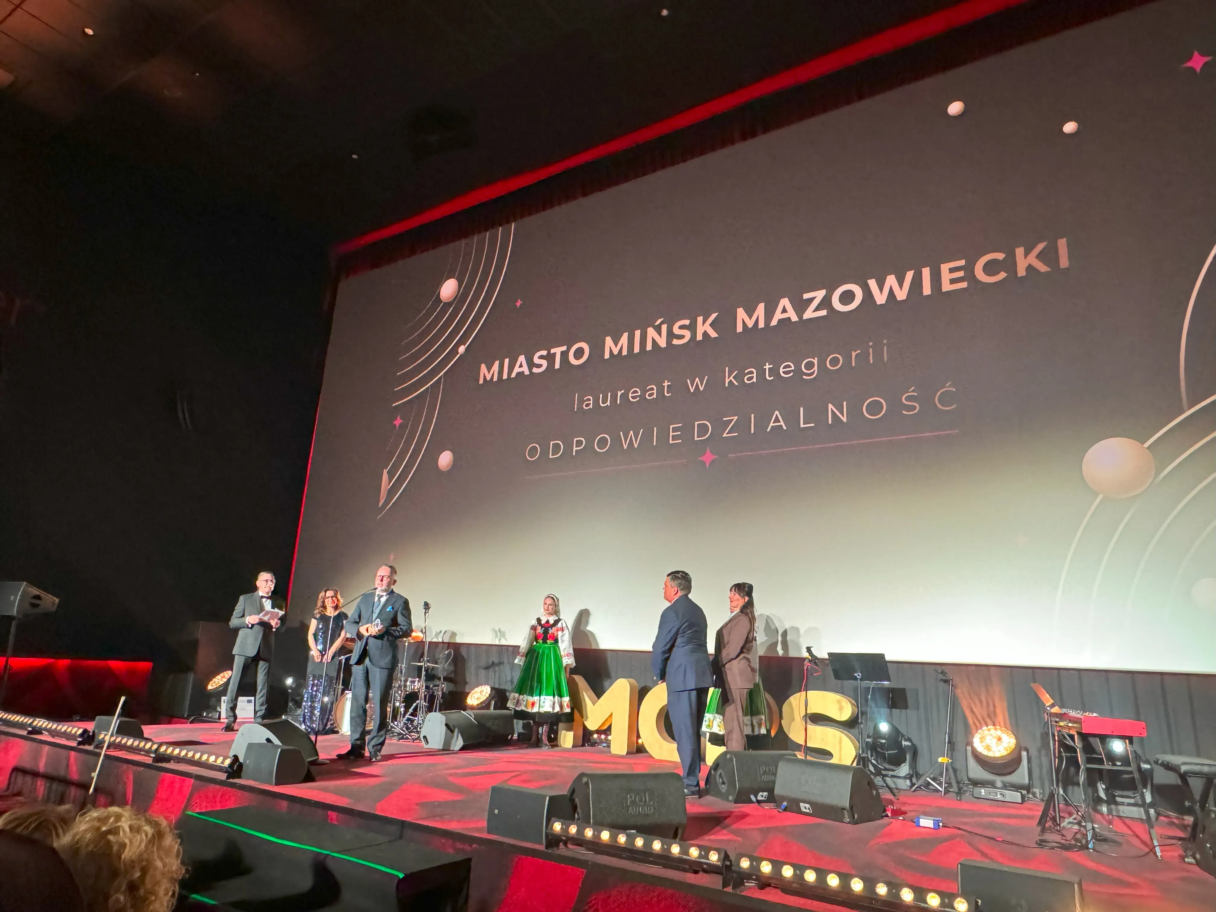 Grupa osób stoi na scenie podczas uroczystości. Za nimi ekran z napisem „Miasto Mińsk Mazowiecki laureat w kategorii Odpowiedzialność”.