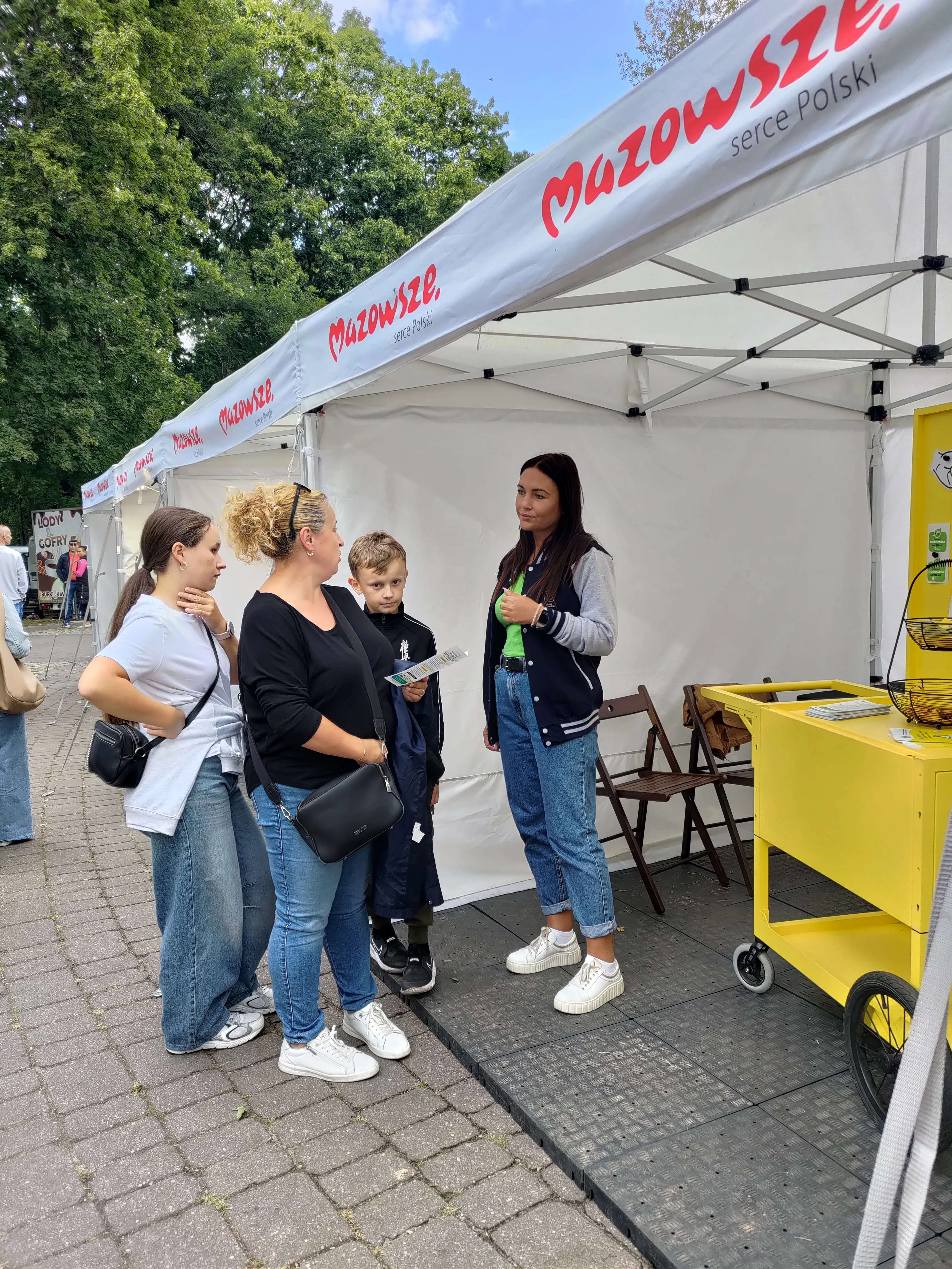 Cztery osoby rozmawiają przy stoisku pod białym namiotem z napisem Mazowsze, na zewnątrz, obok stoi żółty wózek z ulotkami.
