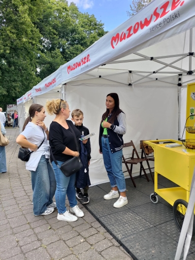 Cztery osoby rozmawiają przy stoisku pod białym namiotem z napisem Mazowsze, na zewnątrz, obok stoi żółty wózek z ulotkami.