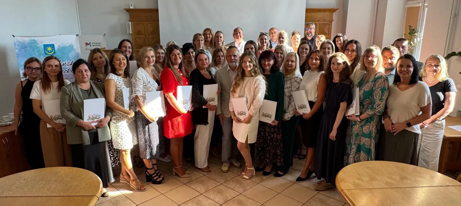 Grupa około 35 osób pozuje do zdjęcia. Stoją w pomieszczeniu, większość trzyma teczki. W tle banery z napisami.